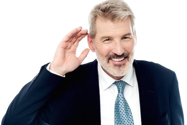older business man in suit holding his hand up to his ear as if he can't hear or wants to hear
