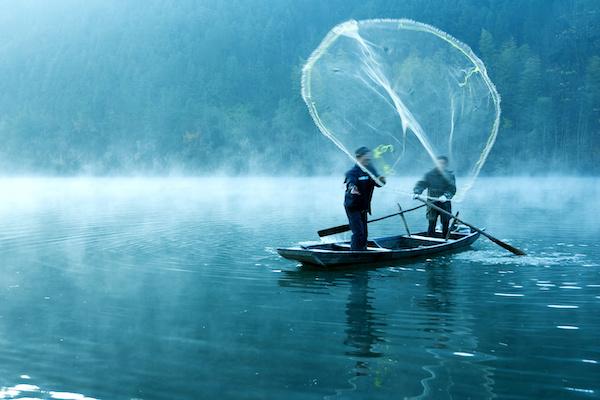 two fishermen in a boat casting a wide net into the water