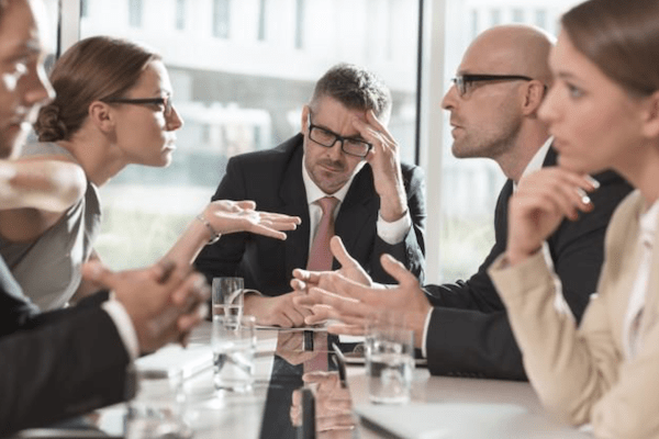 professionals arguing across conference table with man at the head of it looking defeated and discouraged